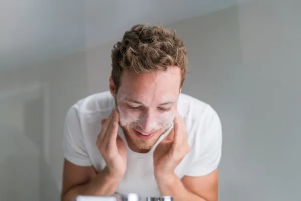 Junger Mann lehnt über ein Waschbecken und reinigt mit Reinigungsschaum sein Gesicht